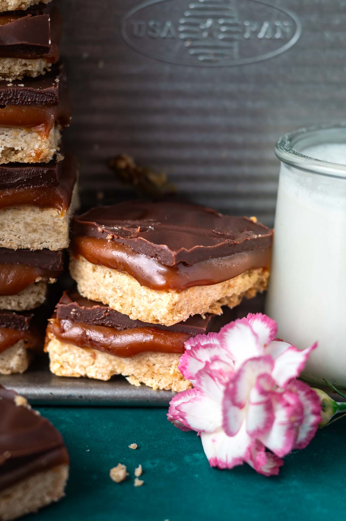 caramel slice bars stacked in a baking pan with a glass of milk and pink flower