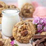 cookie leaning up against a glass of milk with fresh flowers and a bowl of candy bars in the foreground