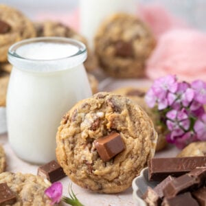 cookie leaning up against a glass of milk with fresh flowers and a bowl of candy bars in the foreground