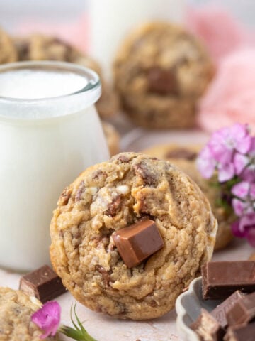cookie leaning up against a glass of milk with fresh flowers and a bowl of candy bars in the foreground