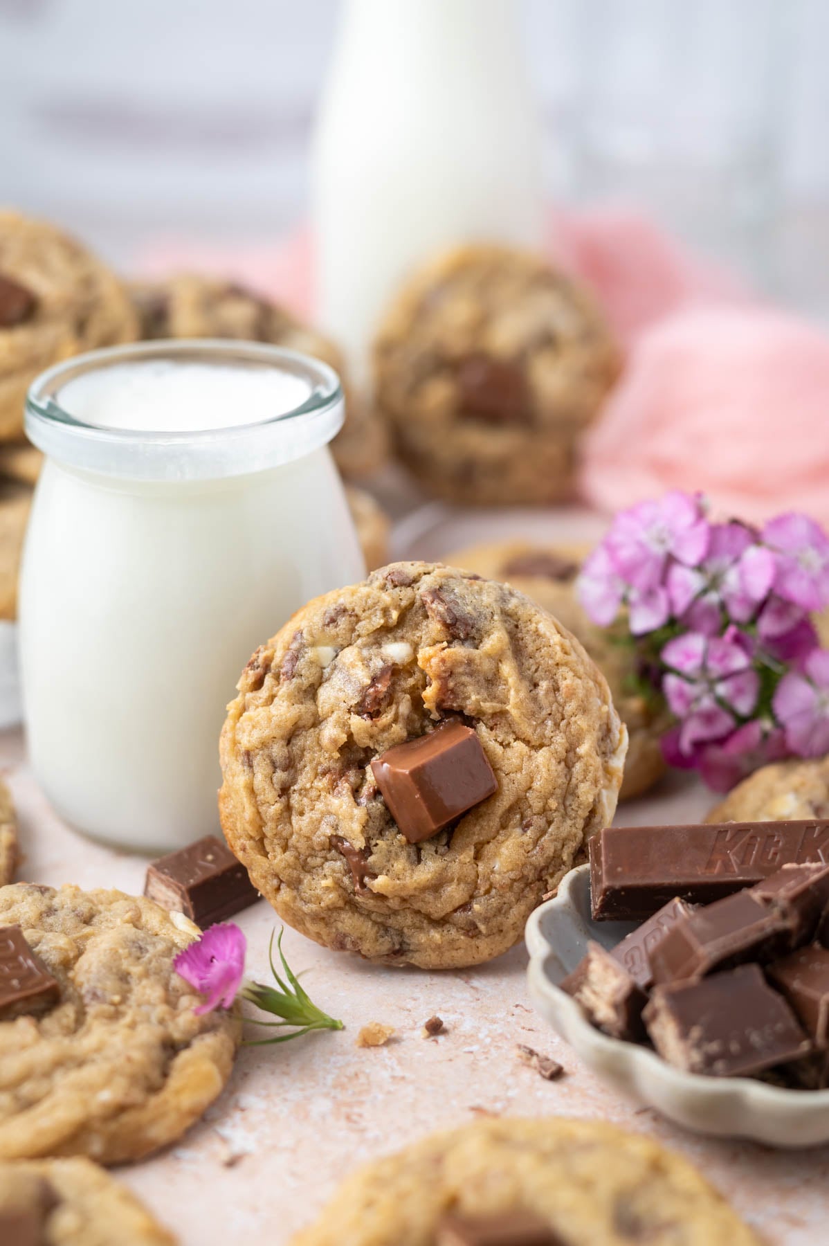 cookie leaning up against a glass of milk with fresh flowers and a bowl of candy bars in the foreground
