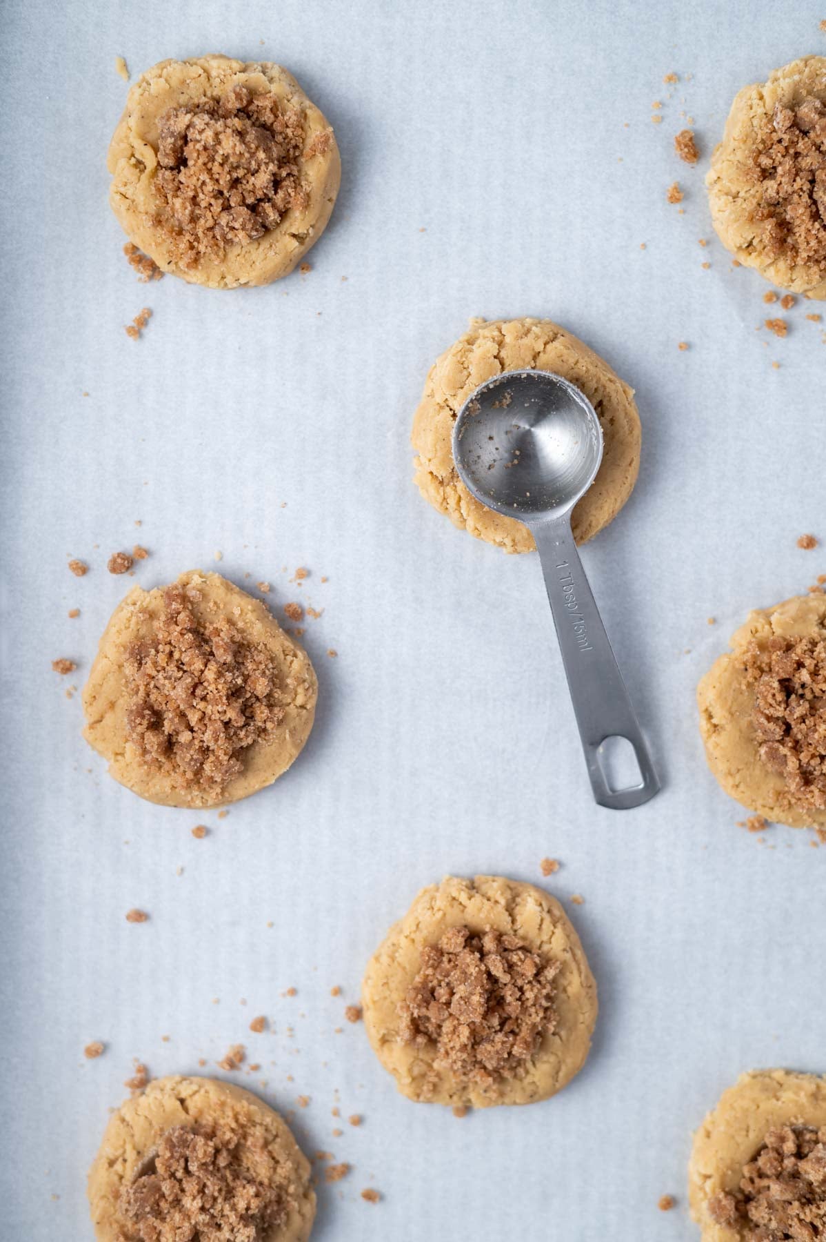 cookie dough filled with streusel on a parchment lined baking sheet with one with a tablespoon pressing the indent