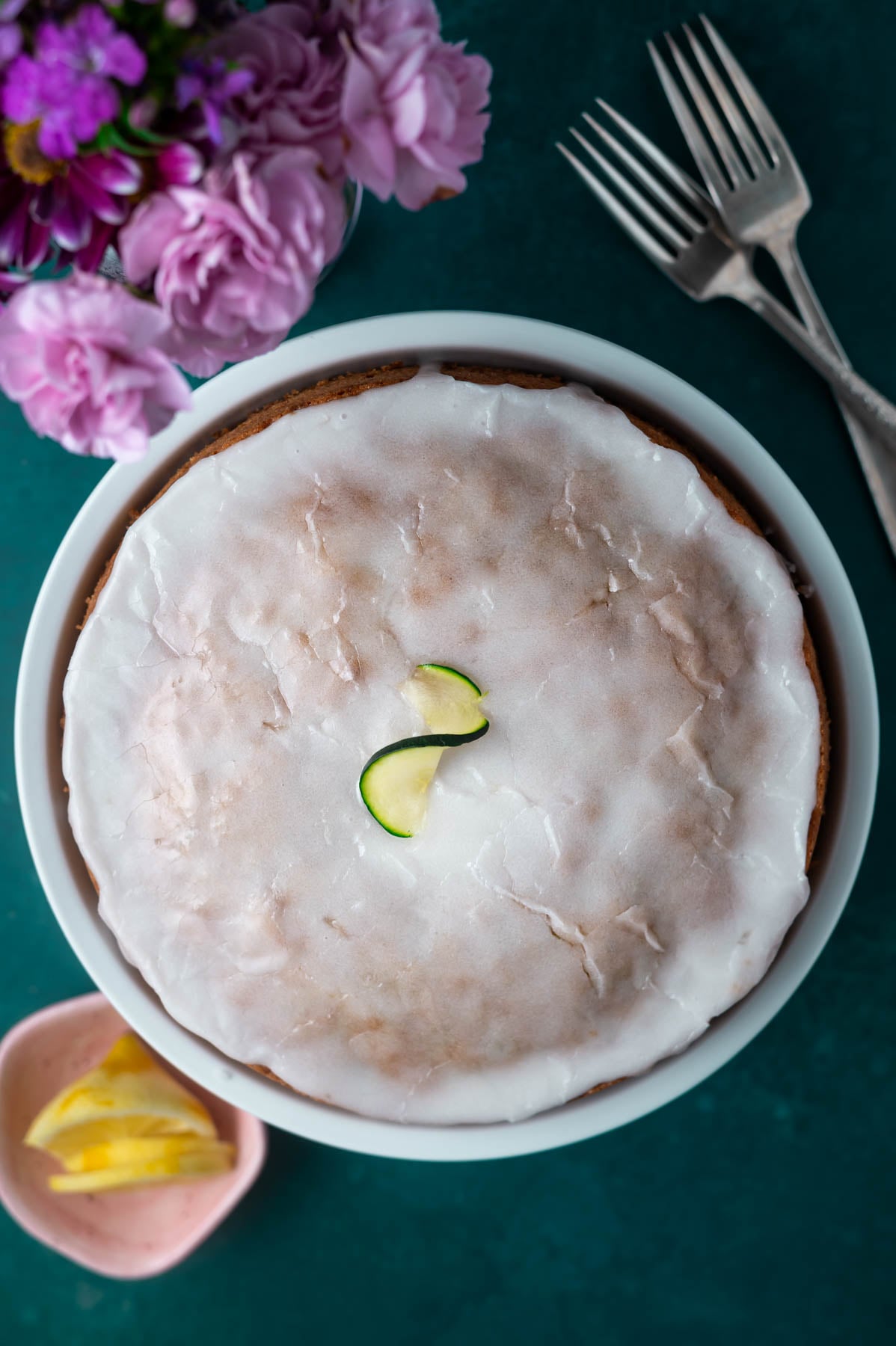 overhead look at a cake on a plate with a lemon glaze and fresh flowers and forks on the side