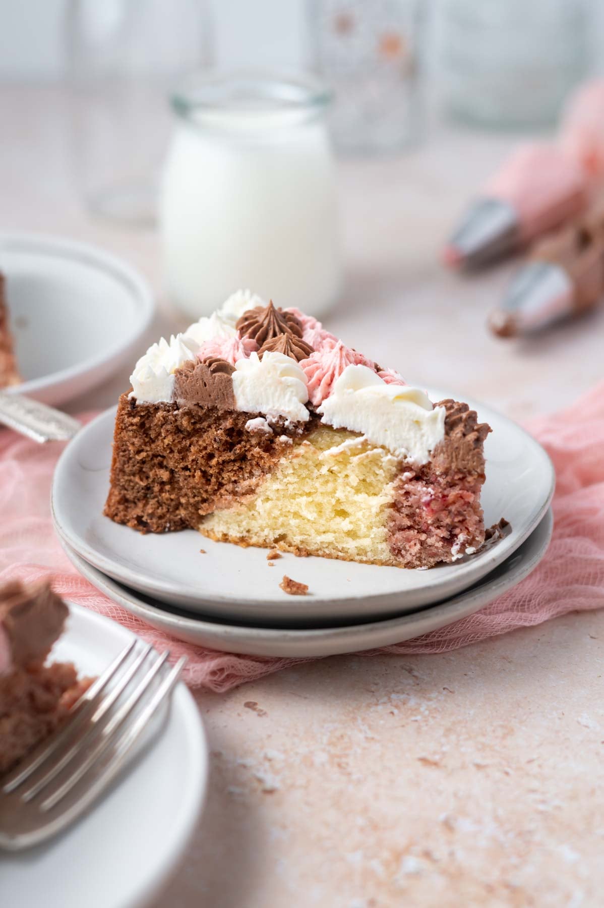 slice of neapolitan cake on a plate with a glass of milk and piping bags of frosting in the background