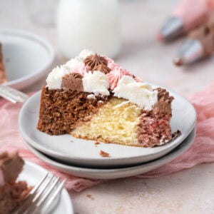 slice of neapolitan cake on a plate with a glass of milk and piping bags of frosting in the background