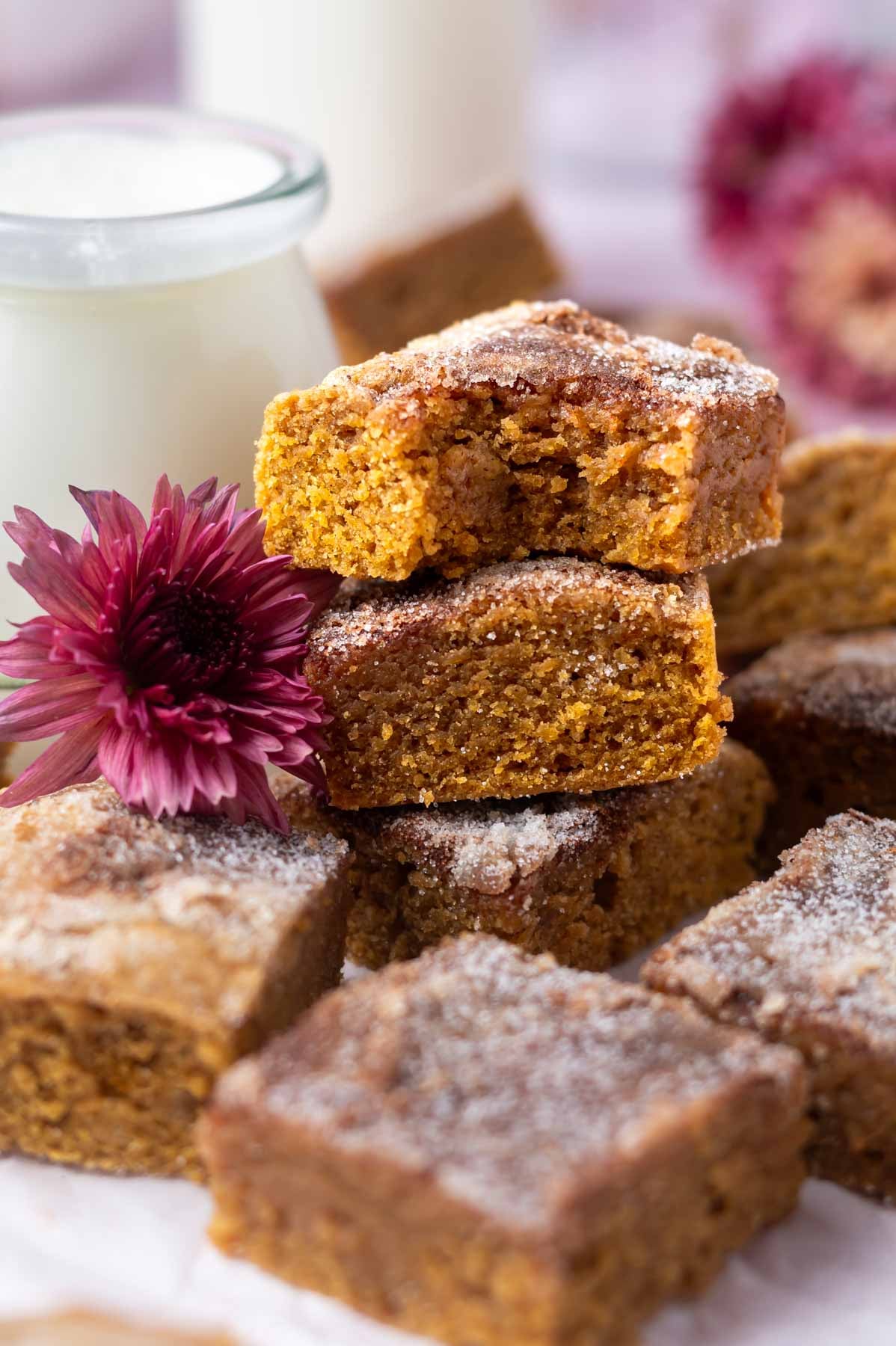 pile of blondie bars with the top one missing a bite and fresh flowers and milk in the background