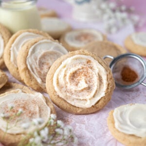 eggnog sugar cookies leaned against a cup of eggnog with a small sifter of cinnamon and nutmeg in the background