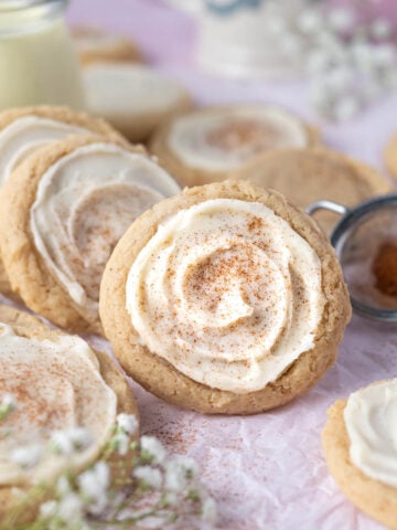 eggnog sugar cookies leaned against a cup of eggnog with a small sifter of cinnamon and nutmeg in the background