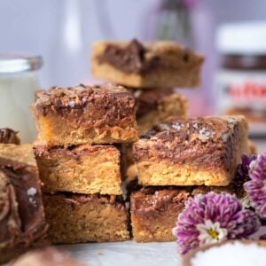 stack of blondies with a glass of milk, jar of Nutella in the background and fresh flowers