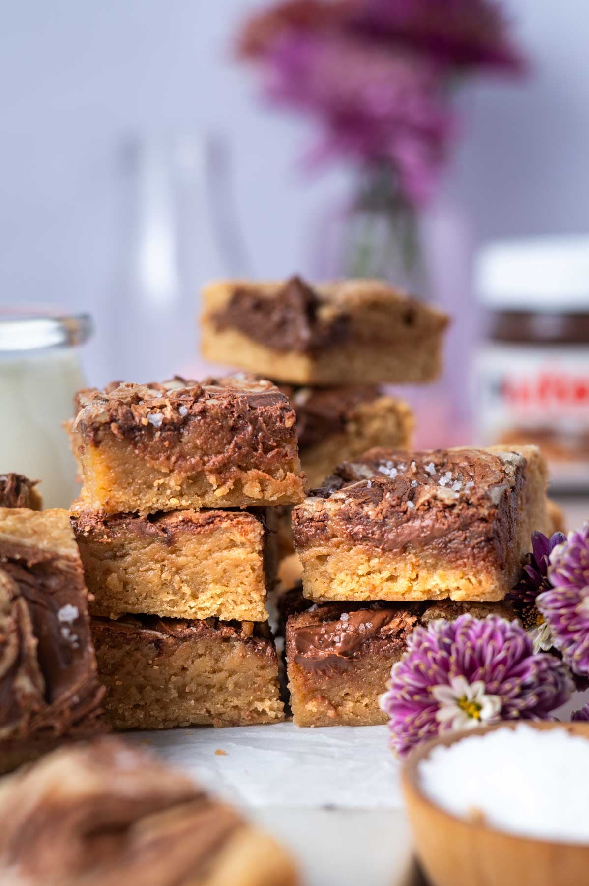stack of blondies with a glass of milk, jar of Nutella in the background and fresh flowers